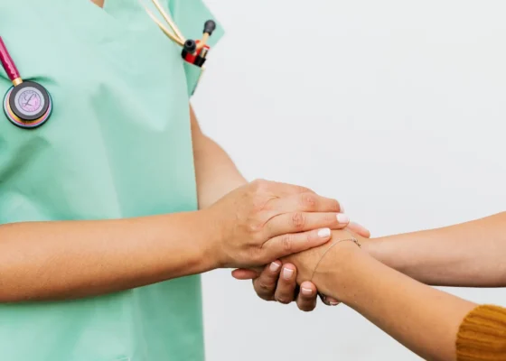 two people clasping hands. Only their arms and hands are visible. We can see one is a medical professional in green scrubs