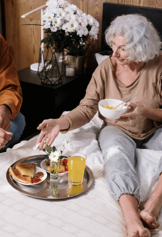 2 people in a bedroom. An older woman is sat on the bed looking at a tray of food. A younger man is sat at the side.