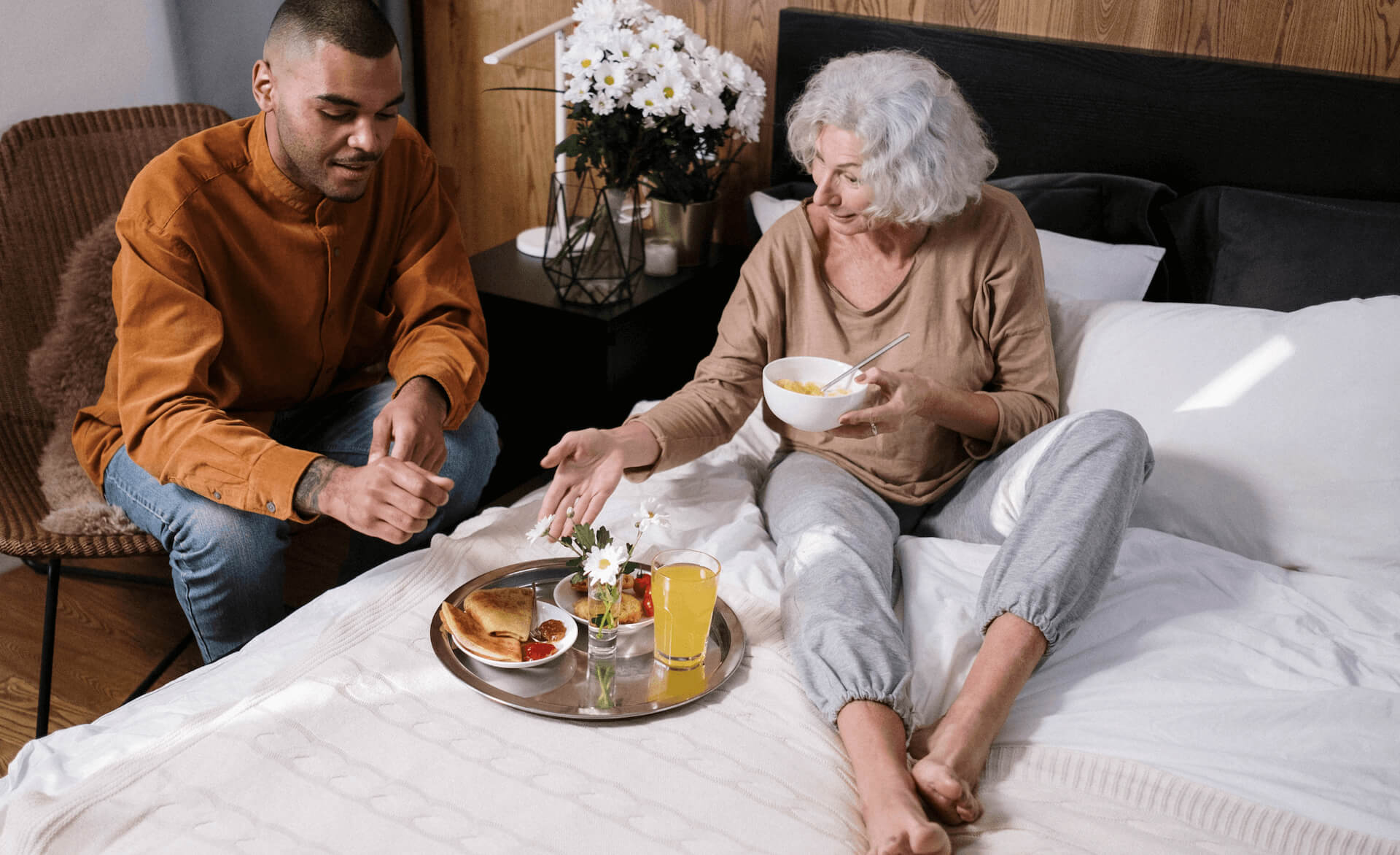2 people in a bedroom. An older woman is sat on the bed looking at a tray of food. A younger man is sat at the side.