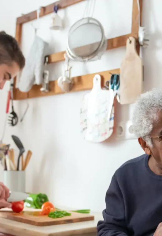 man wearing a navy blue jumper sat at a table but looking over his shoulder. Another man in white is preparing food