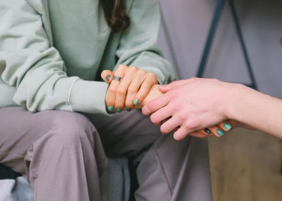 two people sat together. One is wearing hospital scrubs and is holding the hand of the other person
