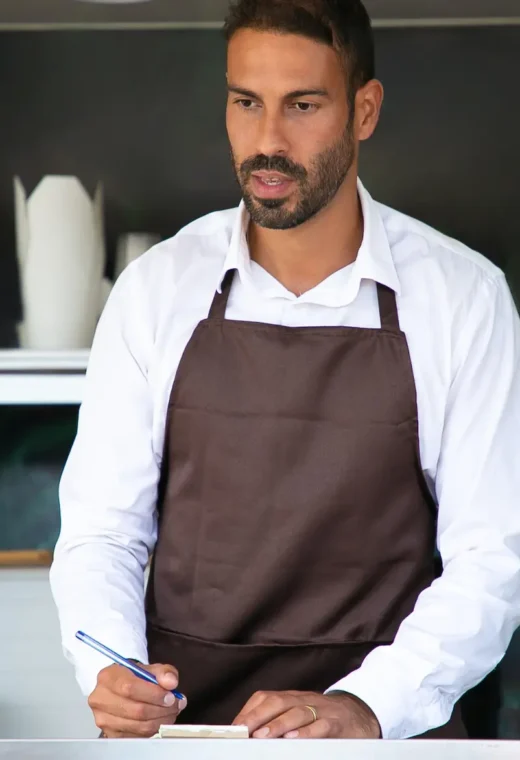 a man wearing chefs whites and a brown apron is holding a pen and writing paper. He is stood at a table.