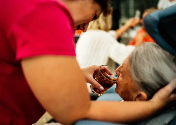 A lady in a red t-shirt helping an older lady take a sip of water.