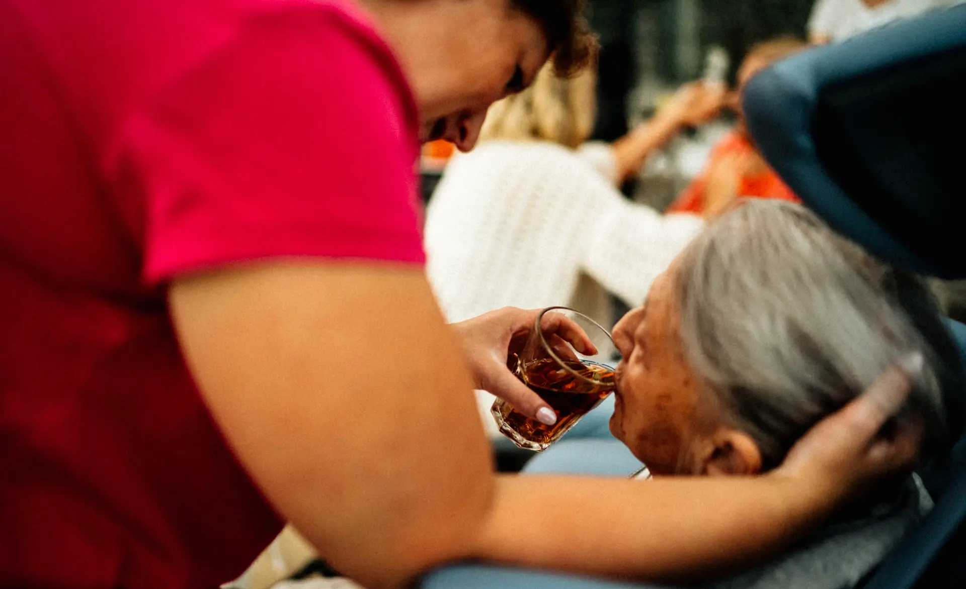 A lady in a red t-shirt helping an older lady take a sip of water.