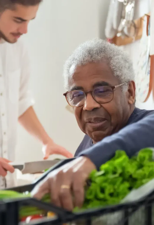 man wearing a navy jumper is holding some leafy green vegetable whilst another man prepares food.