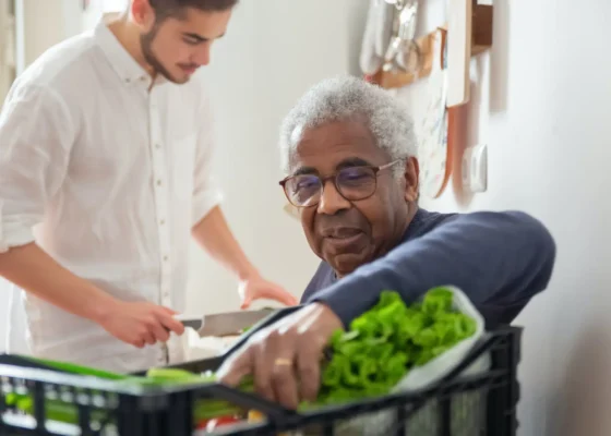 man wearing a navy jumper is holding some leafy green vegetable whilst another man prepares food.