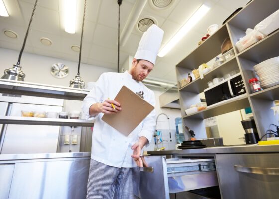 A chef with a clipboard looking in drawers in a kitchen for do agency chefs need a dbs check